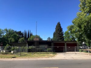 Image of ranch-style building surrounded by a cyclone fence and tall weeds.