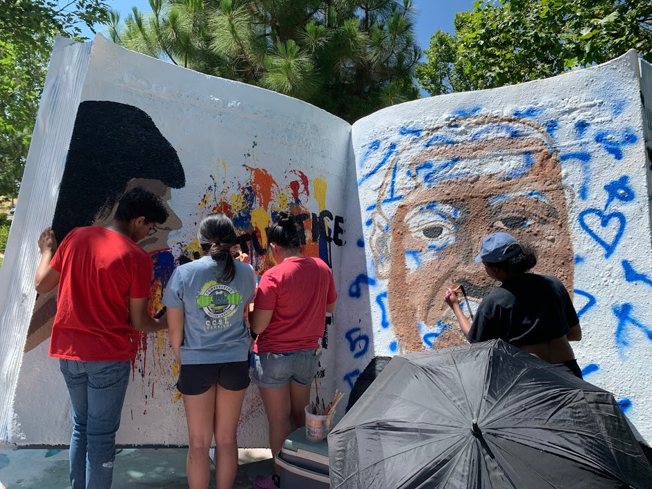 Image of large book. Painted on the left is a person with multiple colors of paint spilling out of their mouth and the words "No Justice No Peace No Racist Police" and painted on the right is a portrait of George Floyd who was killed while in police custody in Minnesota.