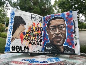 Image of large book. Painted on the left is a person with multiple colors of paint spilling out of their mouth and the words "No Justice No Peace No Racist Police" and painted on the right is a portrait of George Floyd who was killed while in police custody in Minnesota.