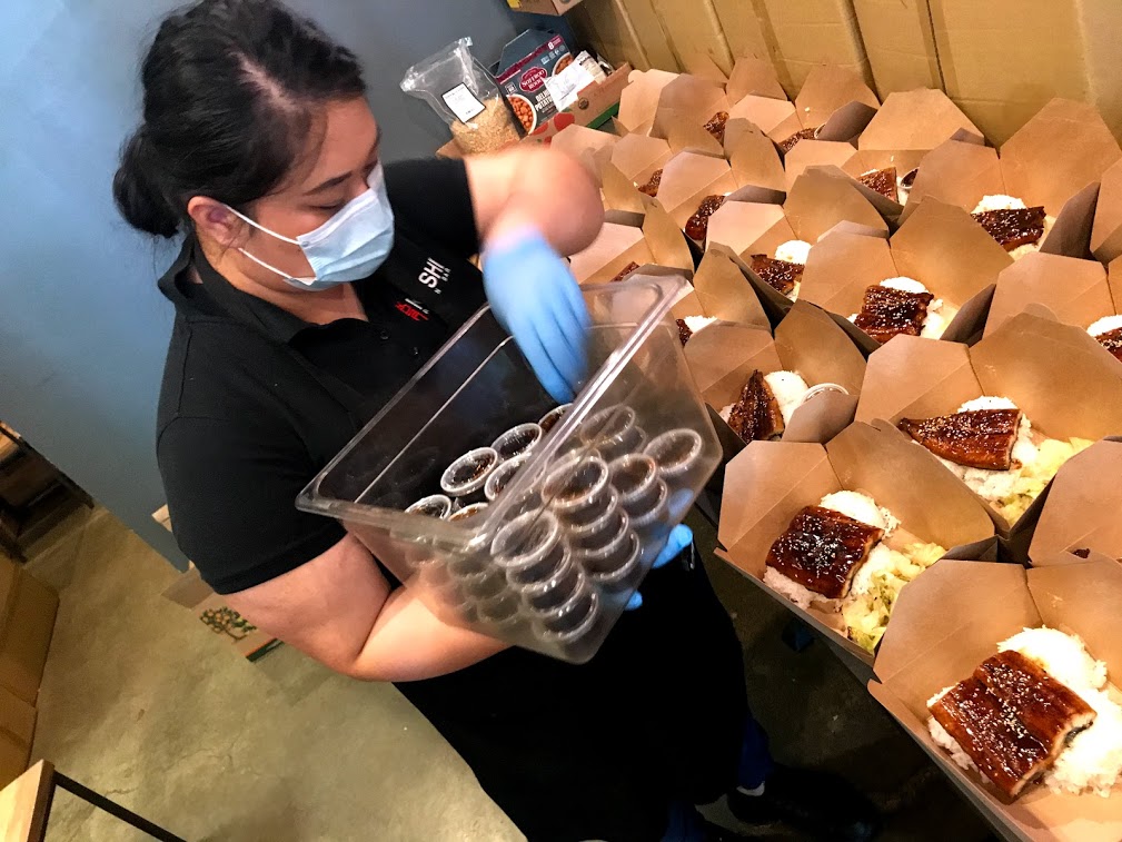 Image of woman wearing face mask and gloves placing small containers of sauce into a boxed meal.