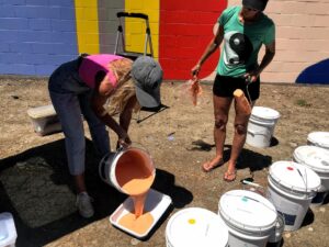 Image of woman pouring paint into a painter's tray while a second woman holds a paint roller nearby.