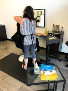 Image of small table with hand sanitizer pump, box of masks and box of gloves in foreground and stylist working on a customer in the background.