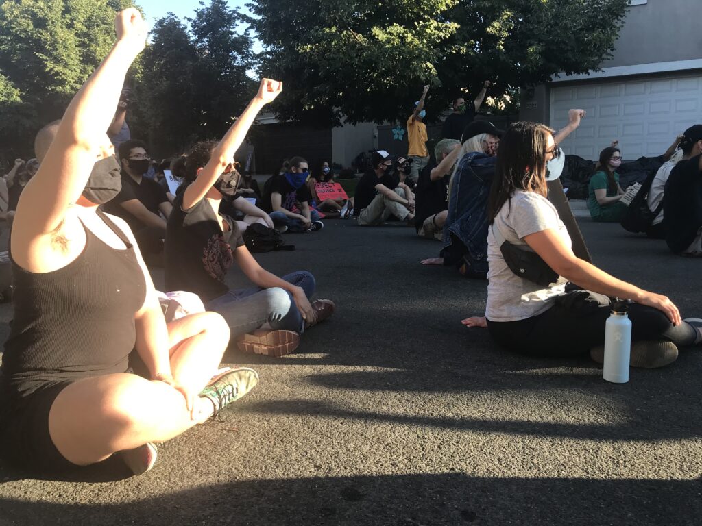 Image of three people in foreground all siting cross legged and holding their fists in the air.