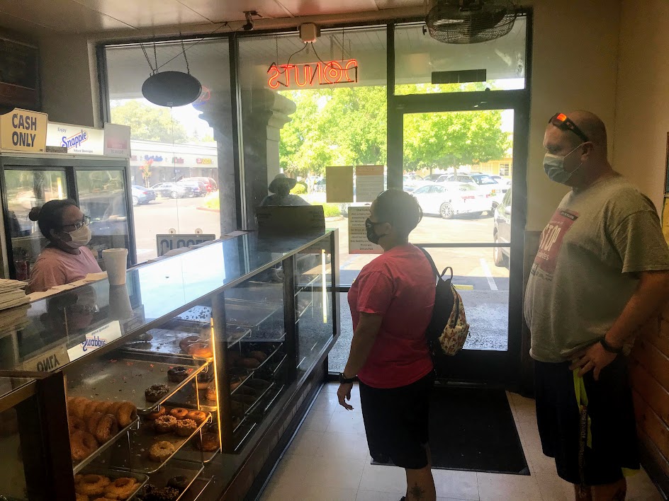 Image of three people talking over a pastry case full of donuts. Everyone is wearing a face mask.