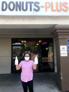 Image of woman standing in front of donuts shop holding up two white paper bags. She is wearing a mask.