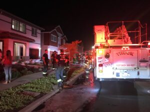 Image of firetruck parked in front of home. It is dark so the truck's lights cast a reddish tint on the building.