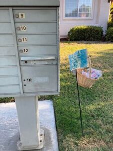 Image of a basket of masks hanging from a post in the ground adjacent to a cluster mailbox.