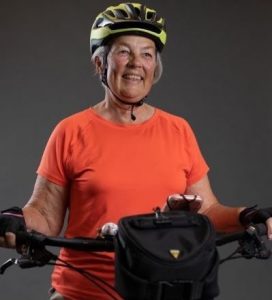 Image of older woman wearing orange shirt and bike helmet standing at bike handlebars.