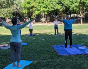 Image of people standing on yoga mats on the grass with arms stretched up and outward.