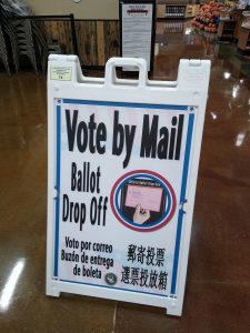 Image of a sign which reads "Vote by Mail" with a ballot drop off box visible in the background.