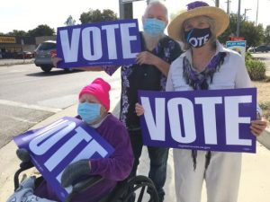 Image of three people holding purple signs with white lettering which reads "VOTE."