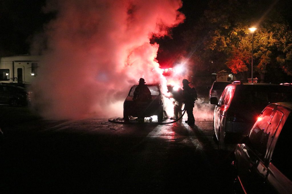 Image of vehicle engulfed in flames. Silhouettes of firefighters visible.