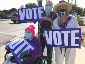 Image of three people holding purple signs that read VOTE. One is in a wheelchair.