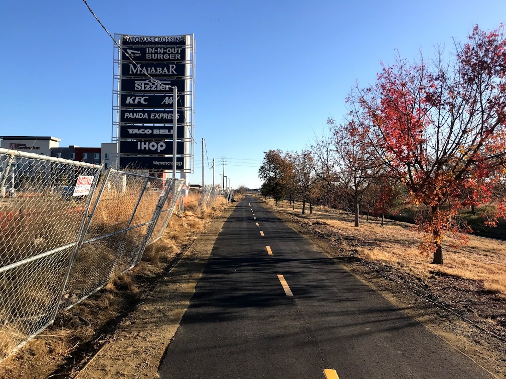 Image of bike trail with trees on the right and a cyclone fence and Natomas Crossing business sign monument on the right. The sign reads In-n-out burger, Malabar, Sizzler, KFC, A&W, Panda express, taco bell, ihop and wienerschnitzel. 