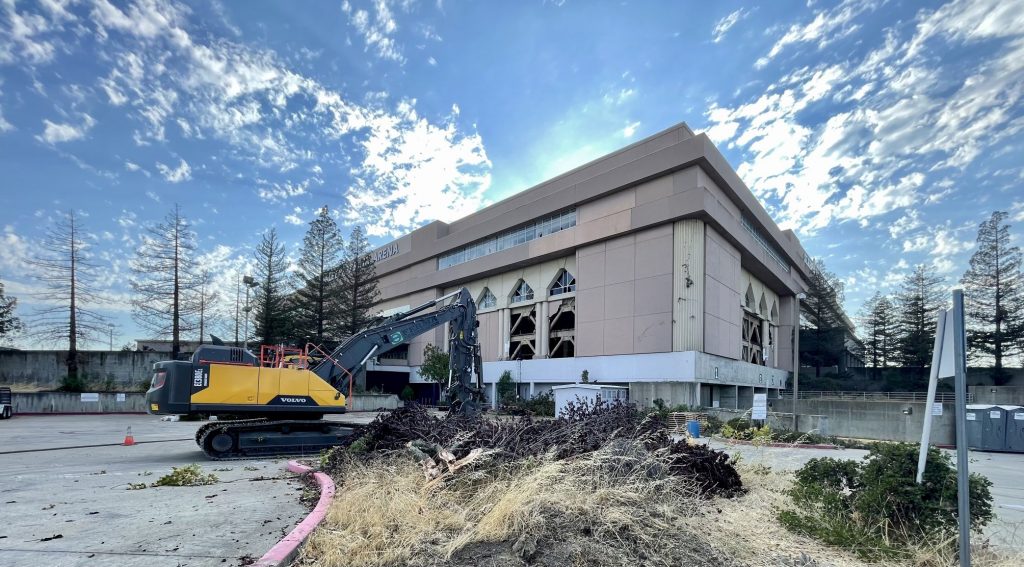 Image of a large backhoe and five-story concrete building. There are open areas where part of the building has been demolished by wrecking crews.