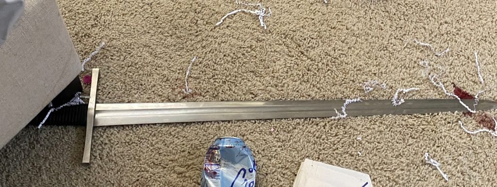 Image of a large, metal sword lying on a carpeted floor. A crushed aluminum can is nearby for perspective.