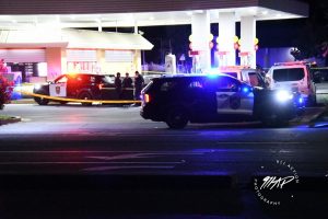 Image of a lighted gas station at night. There are several police cars and yellow crime tape surrounding the area.